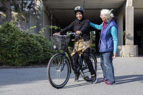 et billede af to kvinder. Den ene sidder på en cykel, og den anden står smilende ved siden af